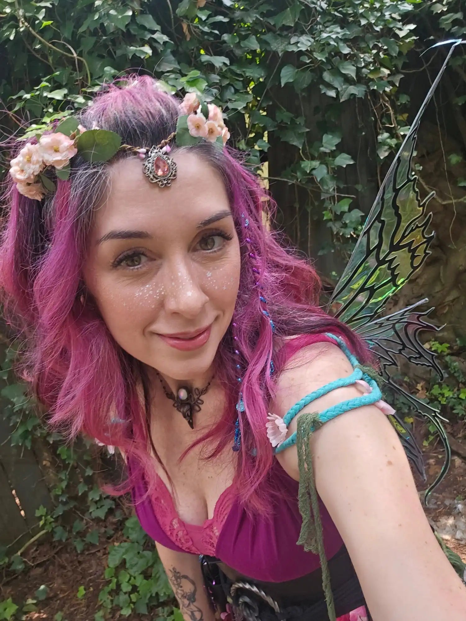 A woman with pink hair, wearing a floral headpiece, a dark choker, a vibrant purple top, and fairy wings.